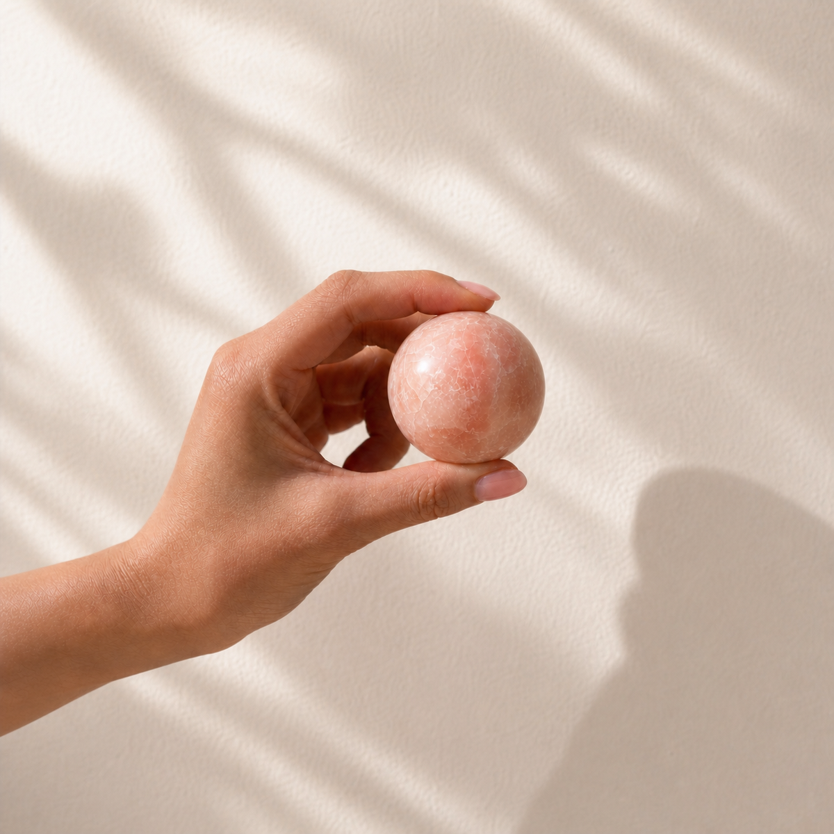 Pink Opal crystal sphere held in hand against a softly lit cream wall to show its size and scale